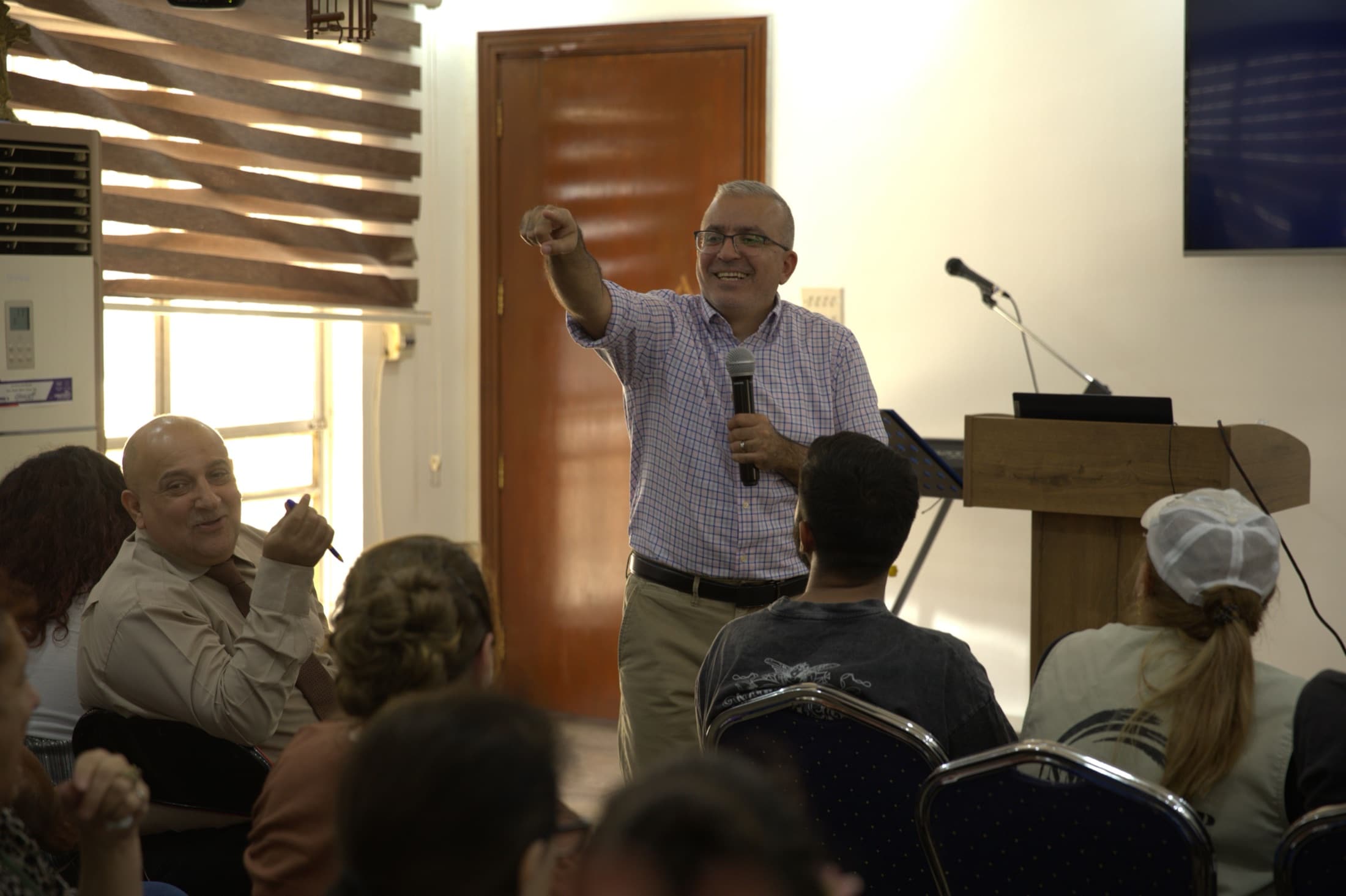 The teacher at the Baghdad hermeneutics training standing among the seated participants, microphone in one hand and pointing toward someone in the room, with the podium and laptop behind him.