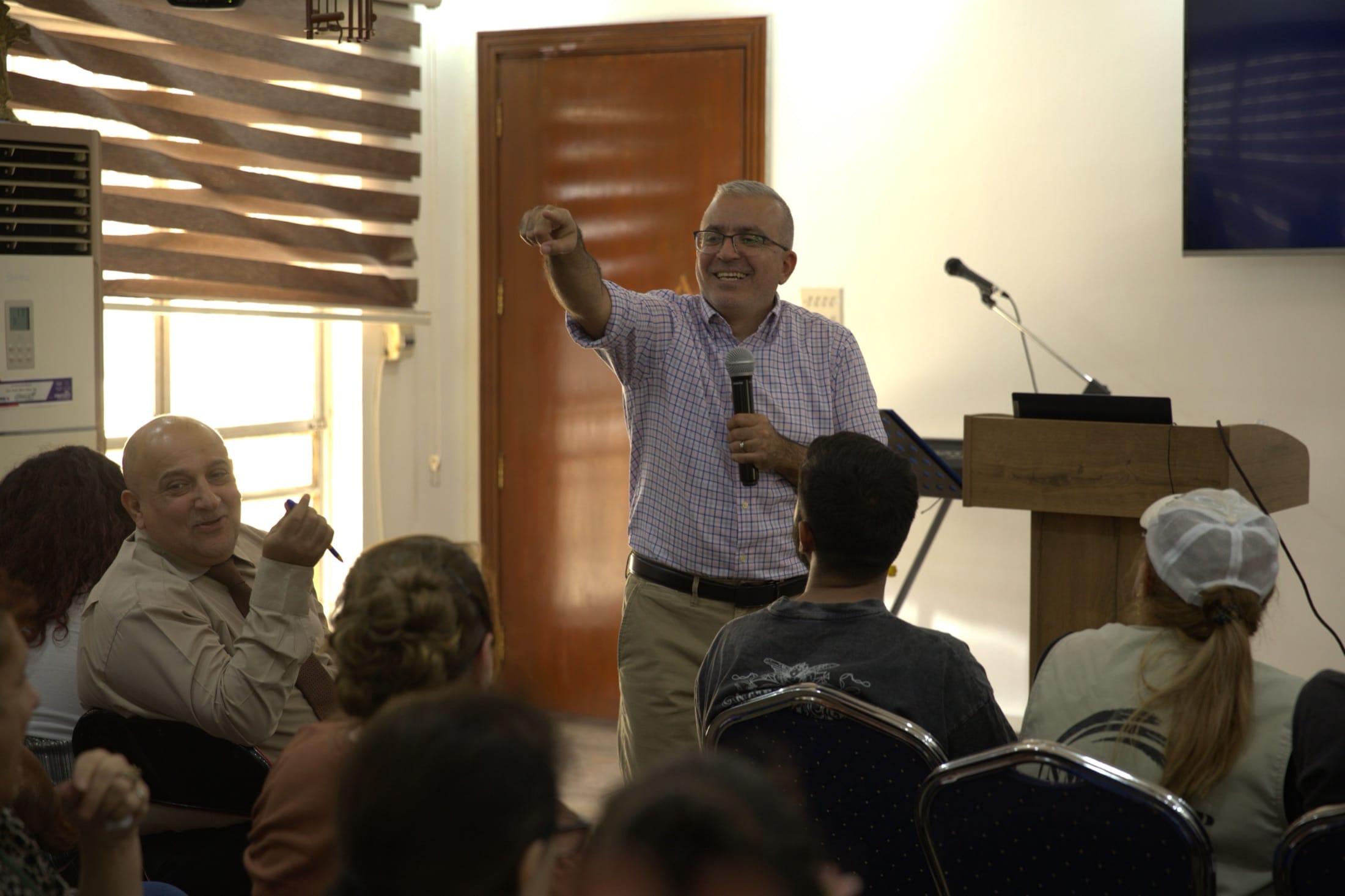 The teacher at the Baghdad hermeneutics training standing among the seated participants, microphone in one hand and pointing toward someone in the room, with the podium and laptop behind him.