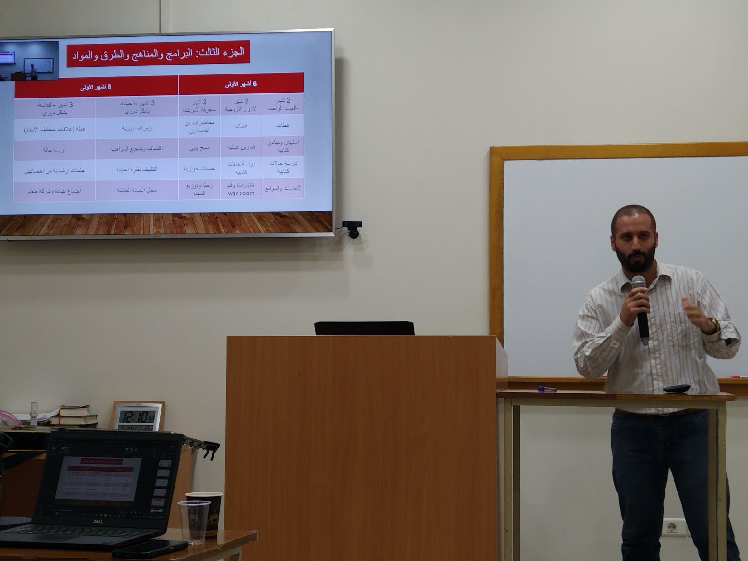 An instructor teaching with a handheld microphone beside a podium and whiteboard, presenting an Arabic curriculum table on a wall-mounted screen.