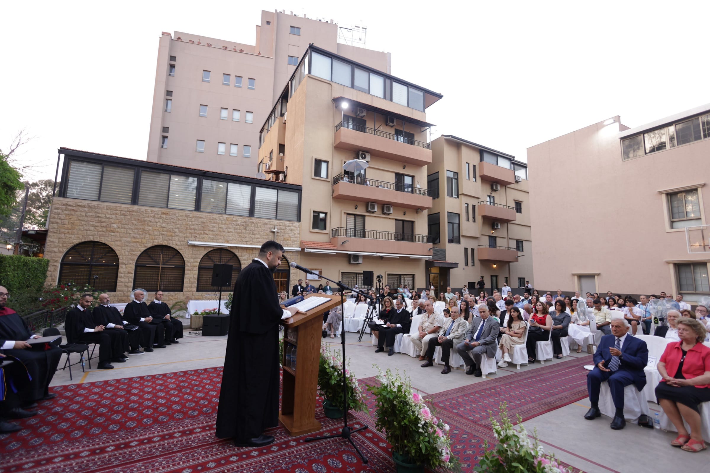 A CAIT graduation service in progress in the courtyard: a robed speaker reading at the podium on the carpeted stage, faculty seated to one side and a large audience to the other, framed by the institute’s stone arcade and the surrounding Beirut apartment blocks.