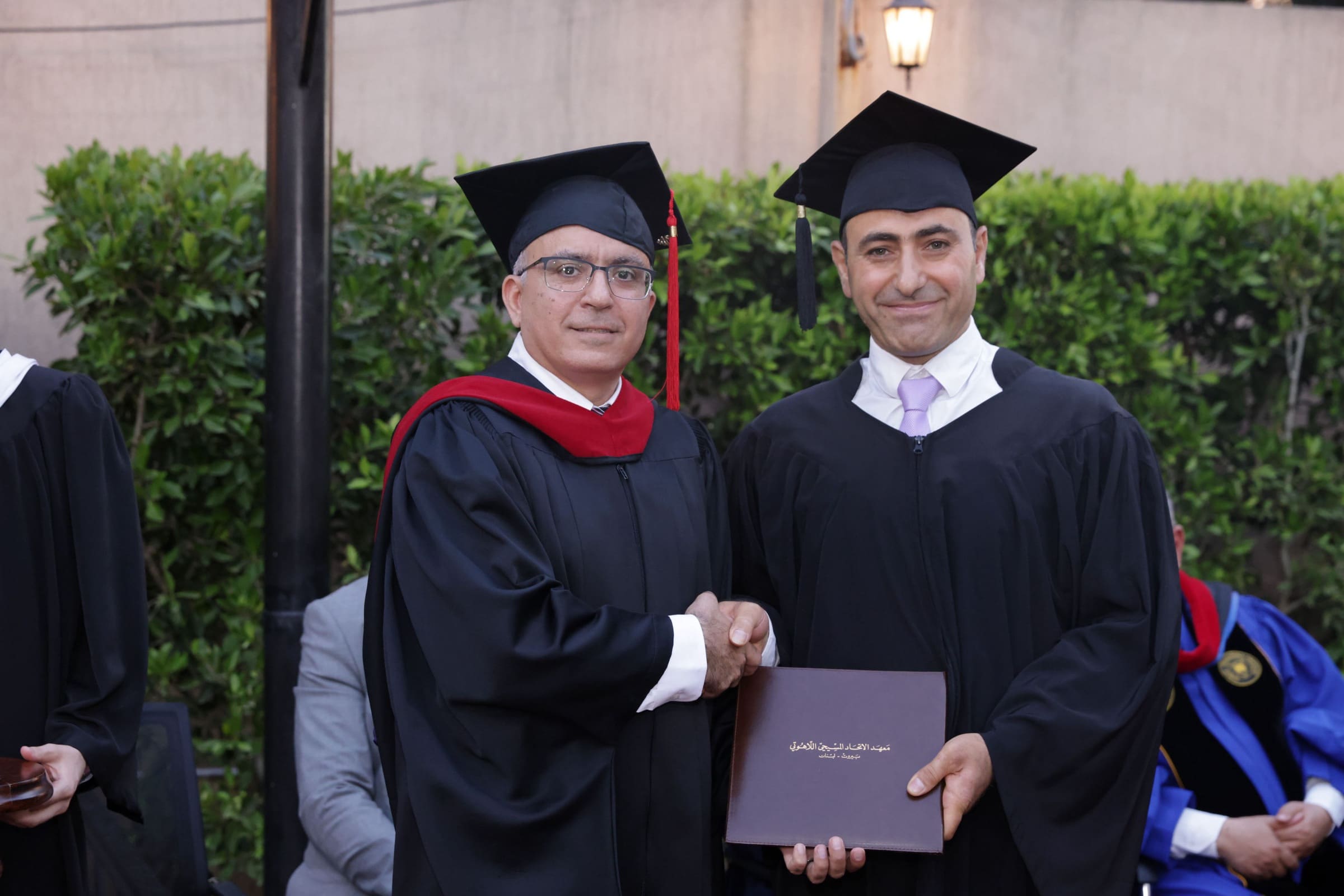 A graduate in cap and gown shaking hands with a hooded faculty member while holding a burgundy diploma folder embossed in Arabic gold foil reading “Christian Alliance Institute of Theology, Beirut – Lebanon”.