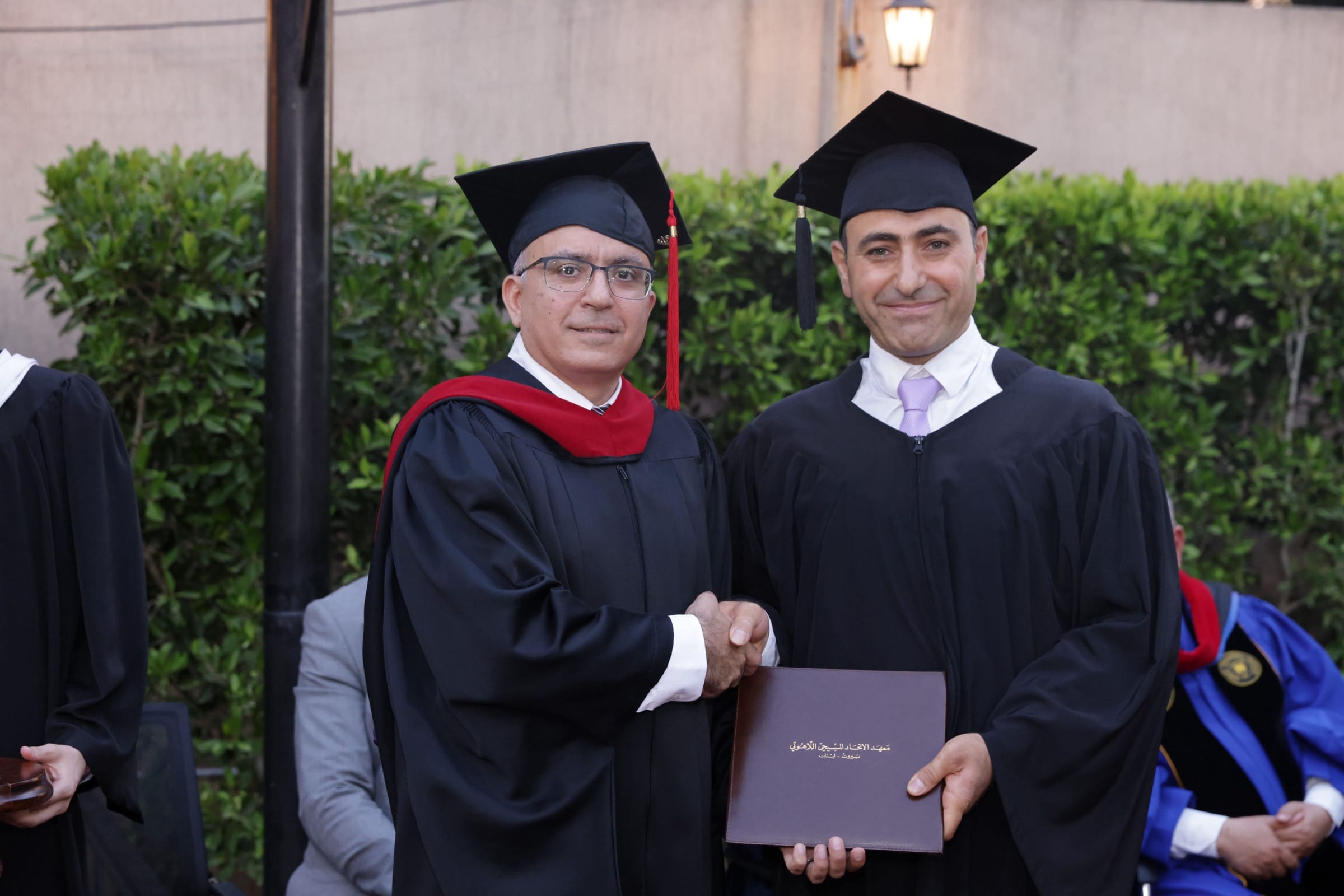 A graduate in cap and gown shaking hands with a hooded faculty member while holding a burgundy diploma folder embossed in Arabic gold foil reading “Christian Alliance Institute of Theology, Beirut – Lebanon”.