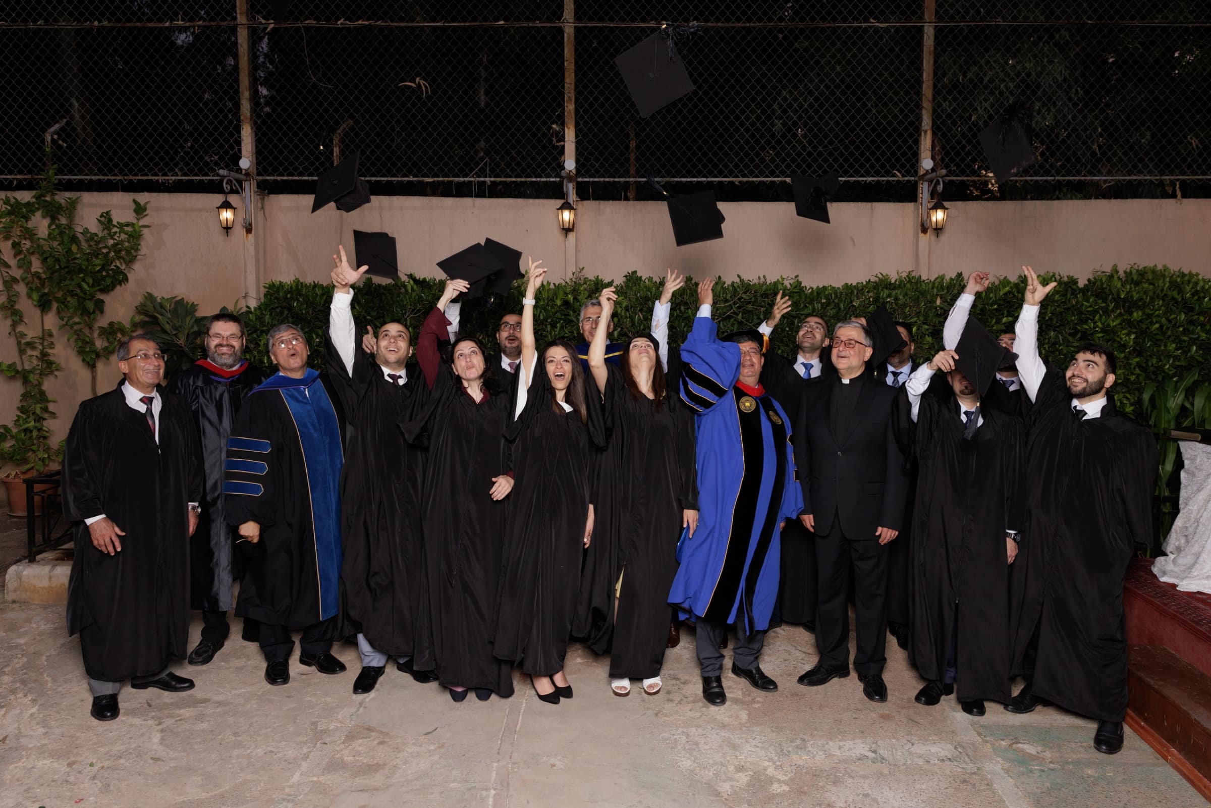 CAIT graduates and faculty in academic gowns throwing their mortarboards into the air in the floodlit courtyard at night, arms raised and a dozen caps caught mid-flight above them.