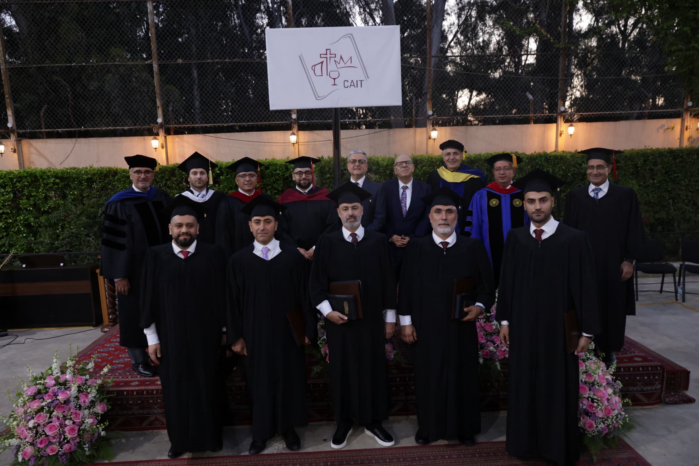 The graduating cohort in caps and gowns standing with faculty and trustees in doctoral regalia on the carpeted courtyard stage, beneath the CAIT banner, for the formal class photograph.