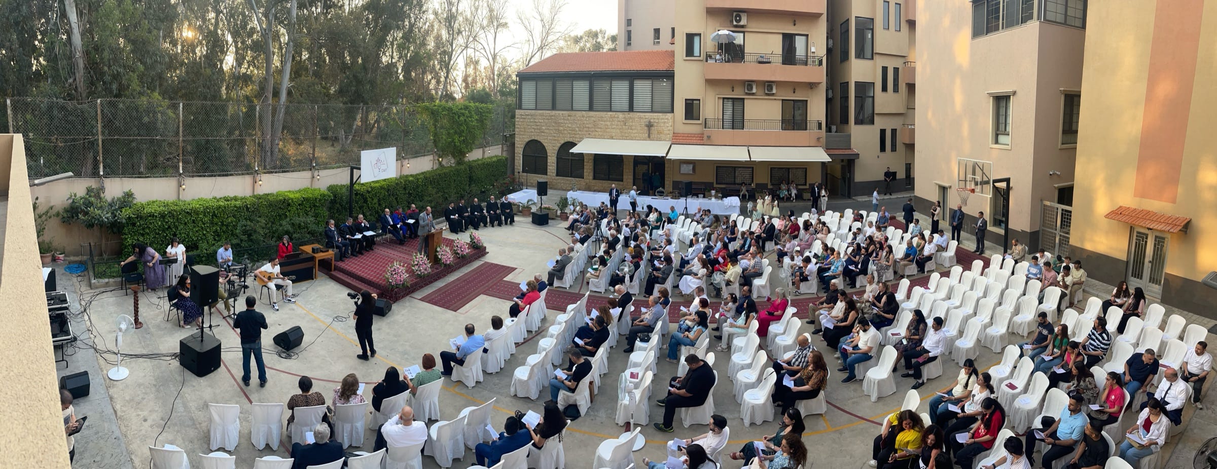 Golden-hour view over the CAIT courtyard during an outdoor graduation service in Beirut: a carpeted stage with faculty seated behind the podium, student musicians leading worship at the left, and a full congregation of families seated on white chairs beneath the surrounding apartment blocks.
