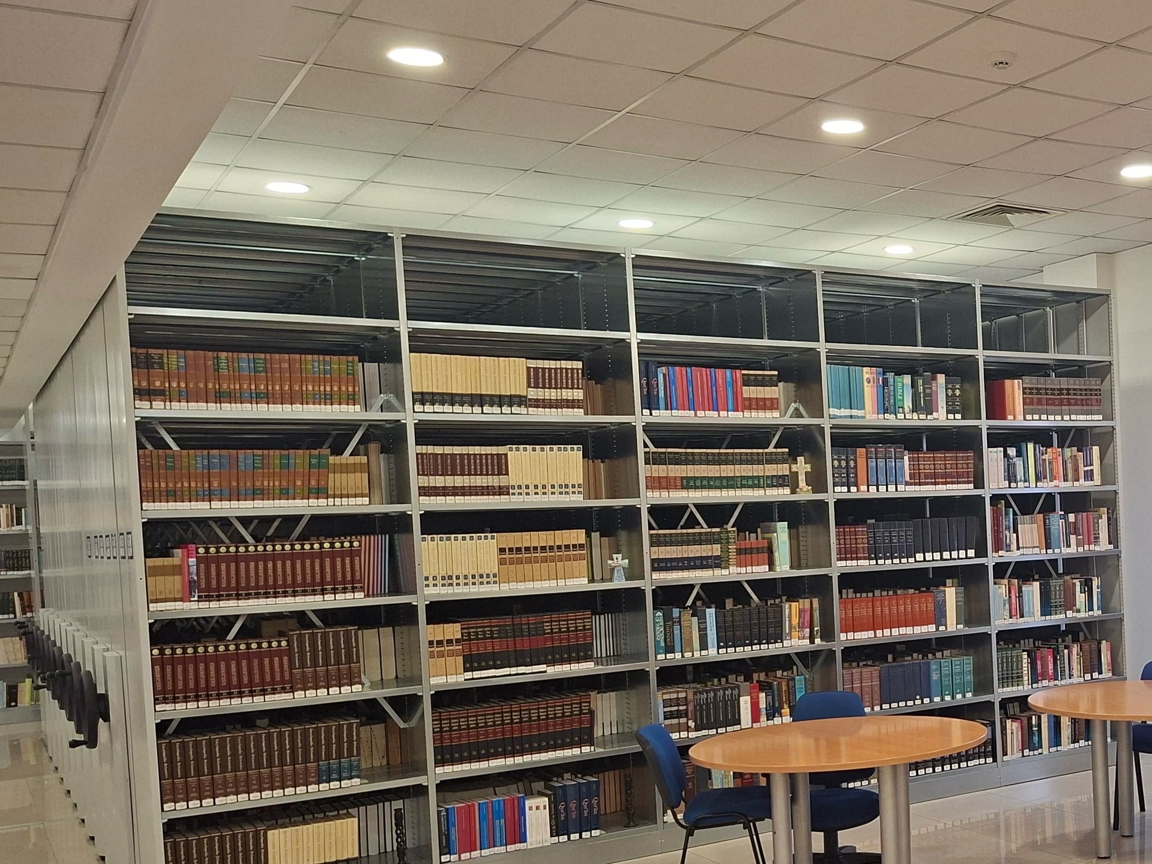The CAIT theological library: a full wall of open shelving packed with commentary sets and reference volumes, with round study tables and blue chairs in the foreground.