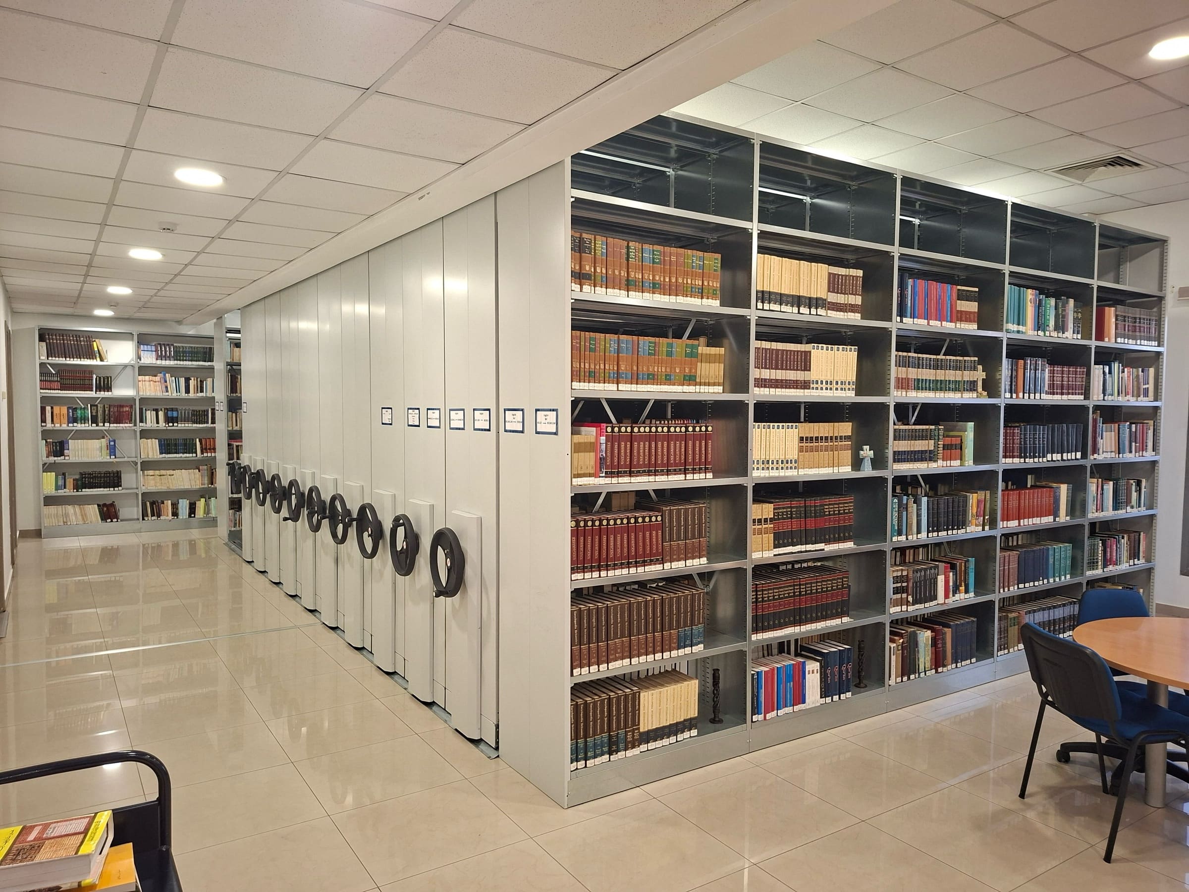 The CAIT theological library: a run of compact rolling stacks with hand-wheel controls, beside fixed shelving filled with theological volumes in Arabic and English, with a round study table and chairs at the right.
