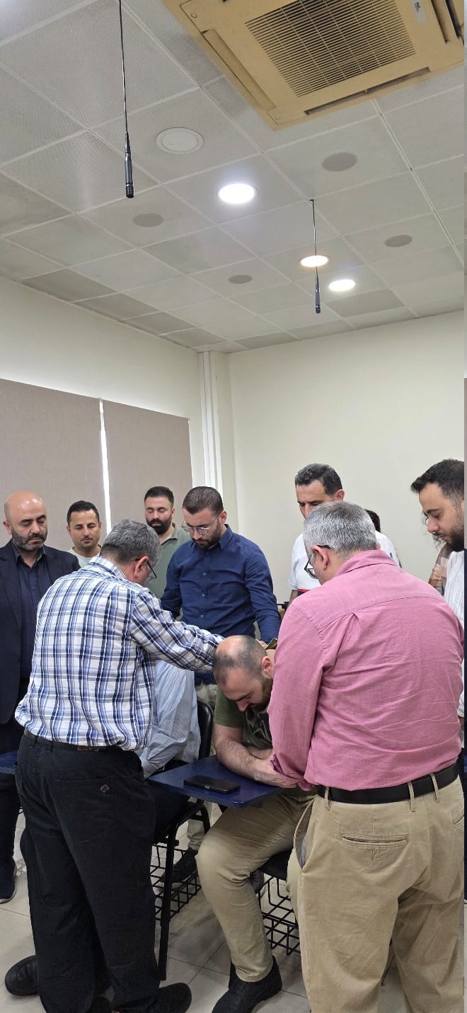 A circle of men standing around a seated man in a CAIT classroom, hands laid on his shoulders and heads bowed, praying over him.