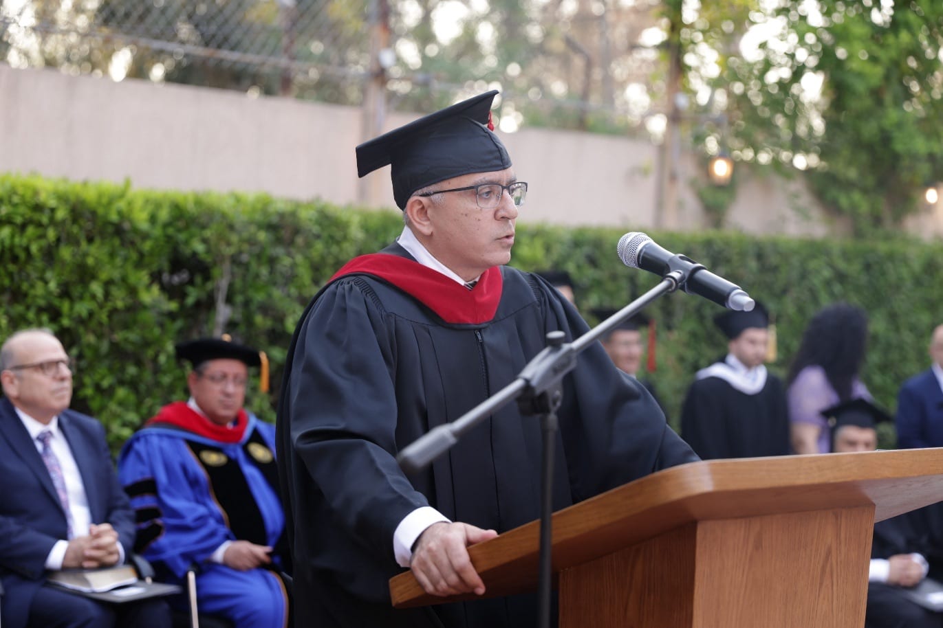Wissam Dagher, President of CAIT, speaking from the wooden podium in cap and gown at the June 2025 graduation, with robed faculty and trustees seated behind him and a hedge and lanterns beyond.