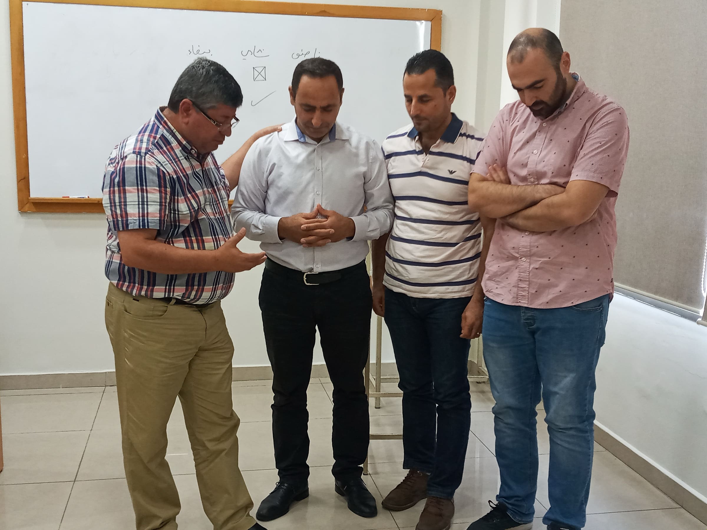 Four CAIT students standing in a circle praying together in a classroom, one with a hand on another’s shoulder, in front of a whiteboard listing Arabic city names.